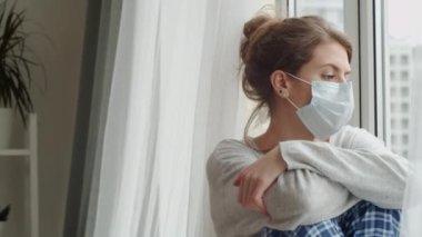 Tıbbi açıdan tek kullanımlık maskeli genç bir kadın karantinada kalırken dizlerine sarılıyor ve şehir caddesinde pencereden dışarı bakıyor.