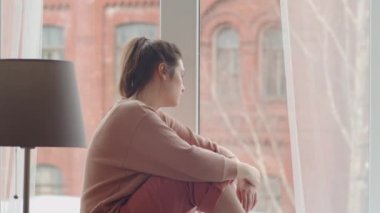 Canı sıkkın genç bir kadın pencerenin kenarına oturmuş, pencereye bakıyor ve evde kilit altında geçirirken parmaklarıyla cama dokunuyor.