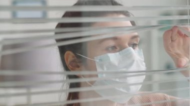 Tıbbi maske takmış genç bir kadının camından panjurları açarken, pencereden bakarken ve karantinada kalırken...