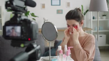 Genç kadın vlogger cilt ürünleri ve ayna ile masada oturuyor, göz bandı takıyor ve evde güzellik kanalı için video kaydederken kameraya konuşuyor