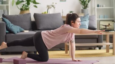 Genç esmer kadın evdeki oturma odasında dizüstü bilgisayarla yoga dersini takip ederken zıt kol ve bacağını spor minderinde uzatıyor.
