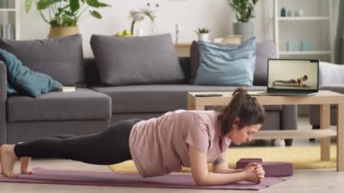 Spor kıyafetleri içinde çıplak ayaklı genç bir kadın kalas egzersizi yapıyor ve sonra dizlerinin üstüne çöküp spor minderinde spor yaparken dizlerinin üstüne çöküp dizlerinin üstüne çöküp dizlerinin üstüne çöküp evde izole bir şekilde antrenman yapıyor.