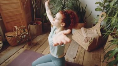 Genç ve güzel bir kadının yeşil bitkilerle odada oturduğu, kollarını kaldırdığı, gözlerini kapattığı ve evde yoga yaparken göğsünün önünde dua pozisyonunda avuç içlerini tuttuğu bir sahne.