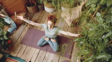 Namaste 'de yeşil tropikal bitkilerle yoga yaparken kollarını kaldırıp göğsünün önünde el ele tutuşan, nilüfer minderinde poz veren genç kızıl saçlı kadının en iyi görüntüsü.