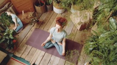 Yeşil bitkilerle dolu bir odada meditasyon yaparken yoga minderinde poz veren ve el ele tutuşan, spor kıyafetli genç kızıl saçlı kadının en iyi görüntüsü.