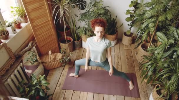 年轻的白种人女子穿着运动服做伸展侧角的运动时在健身垫上摆姿势 同时在有木制地板和绿色植物的房间里练习瑜伽 — 图库视频影像