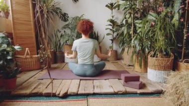 Spor kıyafetli kızıl saçlı kadının spor kıyafetleri içinde nilüfer minderinde poz verirken ve çamurda el ele tutuşurken yoga yaparken ahşap zemin ve yeşil bitkilerle yoga yaparken görüntüsünü büyüt.