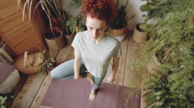 Kızıl saçlı genç bir kadının ağaçta durup Namaste 'de el ele tutuşurken evinde yoga yaparken görüntüsü.