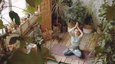 Yüksek açılı bir çekimde Lotus 'ta oturan bir kadın egzersiz minderinde poz verir, dua ederken el ele tutuşur ve sonra yoga yaparken ahşap zemin ve yeşil bitkilerle dolu rahat bir odada meditasyon yapar.