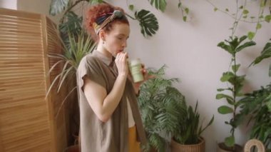 Tekrar kullanılabilen cam samanla çevre dostu bardaktan içen ve sonra yeşil ev bitkileriyle dolu güzel bir odada gülümseyerek kameraya bakan genç bir kadının arkası.