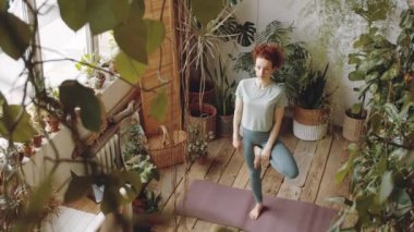 Yüksek açılı çekimde, spor kıyafetli genç bir kadın ağaç duruşu yapıp bir kolunu uzatırken büyük bir odada bir sürü yeşil bitkiyle yoga yaparken görülüyor.