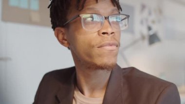 Gözlüklü genç ve yakışıklı Afro-Amerikan işadamının göğüs göğüse çekimleri, sonra kameraya dönüp iş günü masa başında otururken gülümsemeleri.