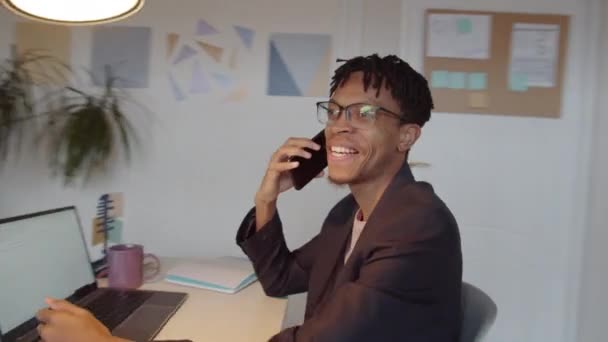 若いアフリカ系アメリカ人ビジネスマンがオフィスのデスクで携帯電話で笑顔で話しているのを360度追跡しながら 彼の女性同僚はバックグラウンドでノートパソコンで作業している — ストック動画