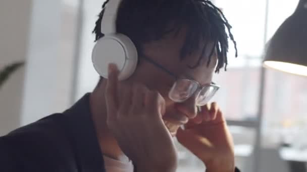 オフィスの彼の職場で無線ヘッドフォンで音楽を聞きながら 彼の頭と笑顔をうなずかせ 若いアフリカ系アメリカ人実業家の360度の追跡ショット — ストック動画