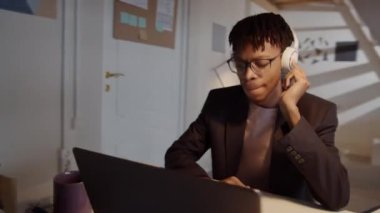 Genç Afrikalı Amerikalı adamın kablosuz kulaklık taktığı 360 derecelik izleme görüntüsü. Ofiste masa başında müzik dinlerken gülümseyip kafasını ritme göre hareket ettiriyor.