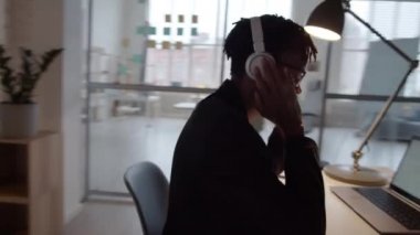 Çalışma masasında müzik dinlerken kablosuz kulaklıklı, gülümseyen ve kafasını ritme göre hareket ettiren genç Afrikalı Amerikalı adamın 360 derecelik dolly çekimi.