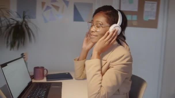 360度ドリーショットの幸せなアフリカ系アメリカ人ビジネスマンの目を閉じて座って オフィスの職場でワイヤレスヘッドフォンで音楽を聴きながらリズムと歌に彼女の頭を移動します — ストック動画