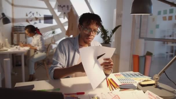 スタジオの作業台に座っている若いアフリカ系アメリカ人男性デザイナーの360度のドーリーショット はさみで紙を切断し カラフルなフェルトの先端ペンでファッションイラストを描く — ストック動画