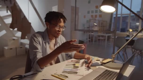 オフィスのデスクでラップトップでオンラインビデオ通話を介して同僚とカラーパレットやファッションイラストを議論するワイヤレスイヤフォンでアフリカ系アメリカ人男性デザイナーの360度ドリーショット — ストック動画