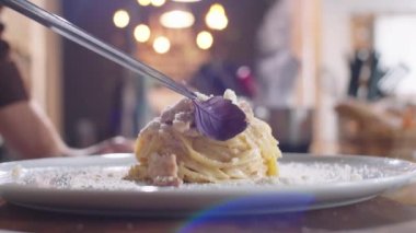 Tabakta servis edilen sıcak carbonara makarnasının üzerine taze fesleğen yaprağı eklemek için mutfak cımbızı kullanan tanınmayan şefin ellerini yakın plan çek.