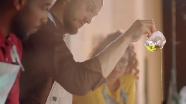 Erkek aşçının tavaya zeytinyağı dökerken çok ırklı öğrencilere yemek pişirme ustası dersi verdiği sahneyi eğ.