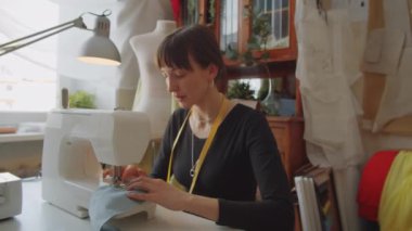 Güzel beyaz kadın atölyede yeni elbiseler üzerinde çalışırken masasında dikiş makinesi kullanıyor.