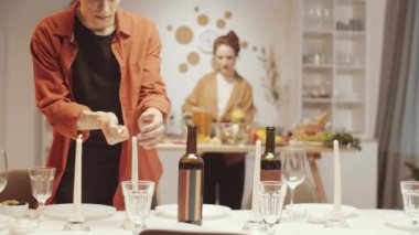 Evli çift evde arkadaşlarını bekliyor: genç beyaz erkek şimşek mumları ve yemek masasını hazırlarken karısı arka planda yemek hazırlıyor.