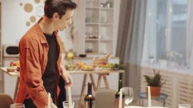 Evde misafir bekleyen genç bir çift: karısı yemek pişirirken neşeli bir adam yemek masasında mum yakıyor.