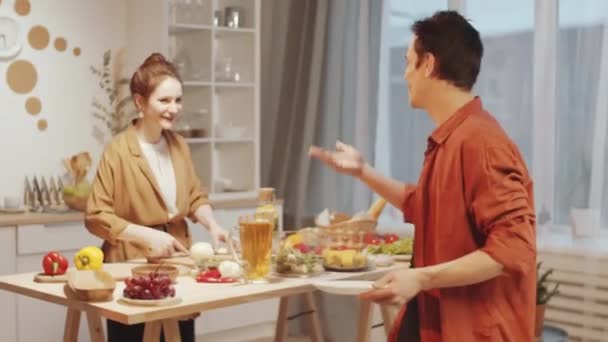 年轻人笑着和快乐的妻子聊天 而妻子则在厨房做饭 然后在准备客人的时候把盘子放在饭桌上 — 图库视频影像