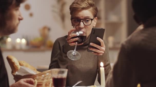 若いです美しいですブロンド女性飲む赤ワインとメッセージ上のスマートフォン彼女の友人ながら何かを議論ホームディナーパーティー — ストック動画