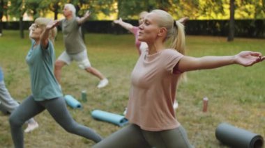 Spor giyim giymiş yaşlı bir grup insanın parkta kadın eğitmenle yoga yaparken hareketlenip kollarını kaldırmasını çek.