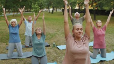 Bir grup yaşlı insan ve kadın eğitmen parkta egzersiz minderleri üzerinde duruyor, kollarını kaldırıyor ve açık havada yoga yaparken Namaste 'ı tutuyorlar.