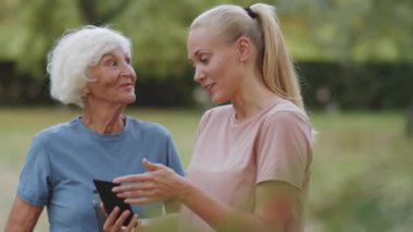 Genç bayan jimnastik koçu, parkta yan yana dururken akıllı telefon ekranında spor giyim konusunda olumlu bir açıklama yapıyor ve gülümsüyor.