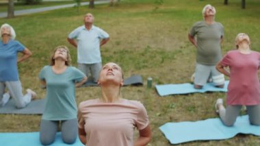 Genç bayan yoga eğitmeni parkta deve pozu verirken yaşlı insanlarla konuşuyor.