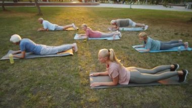 Genç bayan öğretmen ve yaşlı erkek ve kadınlar parktaki minderlerde kobra pozu verirken gün batımında grup yogası yapıyorlar.