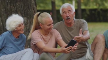 Bayan fitness eğitmeni, spor yaptıktan sonra parkta mola verirken akıllı telefon ekranında yaşlı kadın ve erkeklerle bir şeyler tartışıyor.