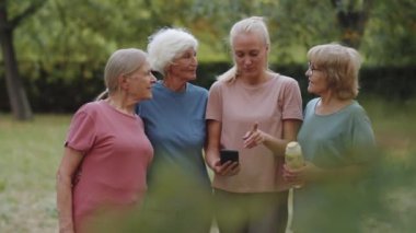 Genç bayan fitness koçu parkta yan yana dururken spor giyim sektöründe yaşlı bir kadınla akıllı telefon ekranında bir şeyler tartışıyor ve gülümsüyor.