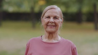 Parkta dikilip kameraya bakıp gülümseyen neşeli, son sınıf öğrencisi bir kadının portresi.