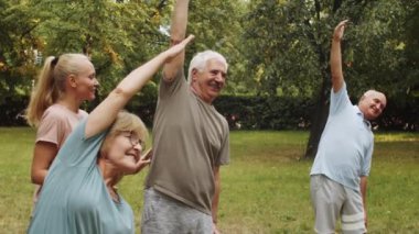 Genç bayan antrenör parkta grup fitness kursu sırasında yan esneme egzersizi yapan neşeli son sınıf öğrencilerine yardım ediyor.