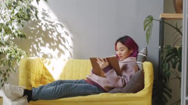 Modern ofisteki kanepede uzanmış kitap okuyan Asyalı bir kadının resmi.