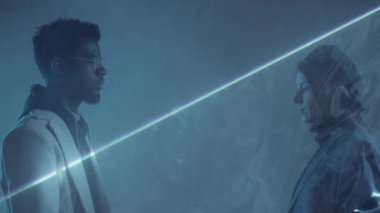 Beyaz tenli genç bir kadının lazer ışığı altında yürüdüğünü ve karanlık stüdyoda sisle Afro-Amerikalı bir adamı kucaklayışını.