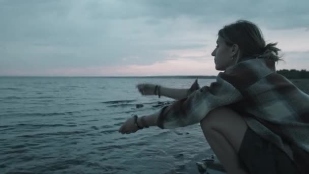 夕方に湖畔に座り 小石を水に投げ入れる若い女性 — ストック動画