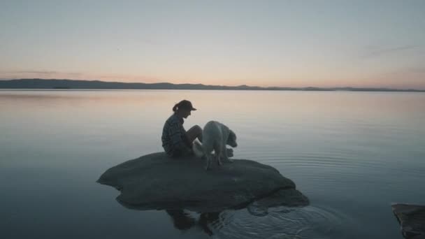 湖の岩の上に座っている女性のシルエットの広いショットと日没時に犬をペッティング — ストック動画