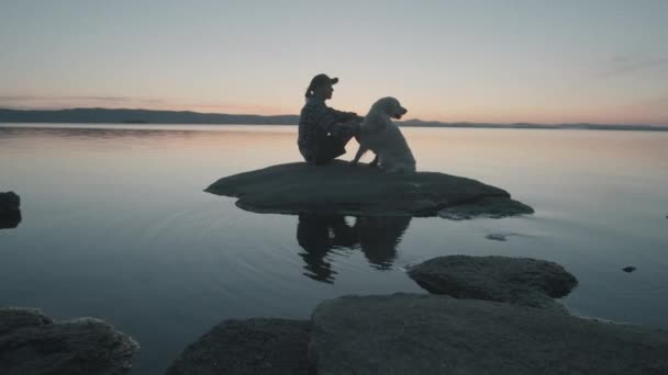 湖の岩の上に座っている女性の広いショット 黄金の回収犬をペットと美しい夕日を見て — ストック動画
