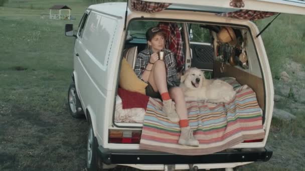 年轻而快乐的女人拿着钢制茶杯 爱抚着可爱的金发碧眼的猎犬 一起坐在大篷车的后门 环视四周的自然 — 图库视频影像