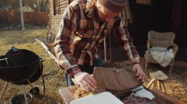 Şapkalı ve oduncu gömlekli bir adam çiftlik evinin arka bahçesinde barbekü yapmaya hazırlanırken bıçakları kontrol ediyor.
