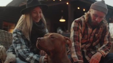 Tapılası pitbull köpeği dışarıda şenlik ateşinin yanında oturuyor ve erkek ve kadın sahipleri tarafından okşanmaktan zevk alıyor.