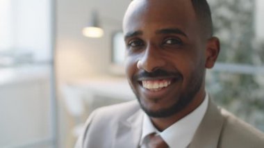 Resmi giyinmiş genç Afro-Amerikalı iş adamının portresi. Kameraya bakıyor ve ofiste otururken gülümsüyor.