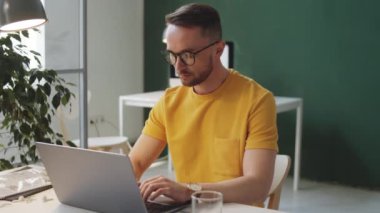 Günlük giysili ve gözlüklü yakışıklı beyaz adam ofisteki laptopta çalışıyor.