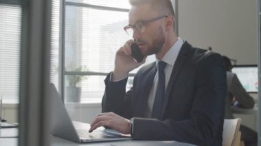 Resmi takım elbiseli ve gözlüklü beyaz işadamı ofis masasında dizüstü bilgisayarla çalışırken cep telefonuyla konuşuyor.